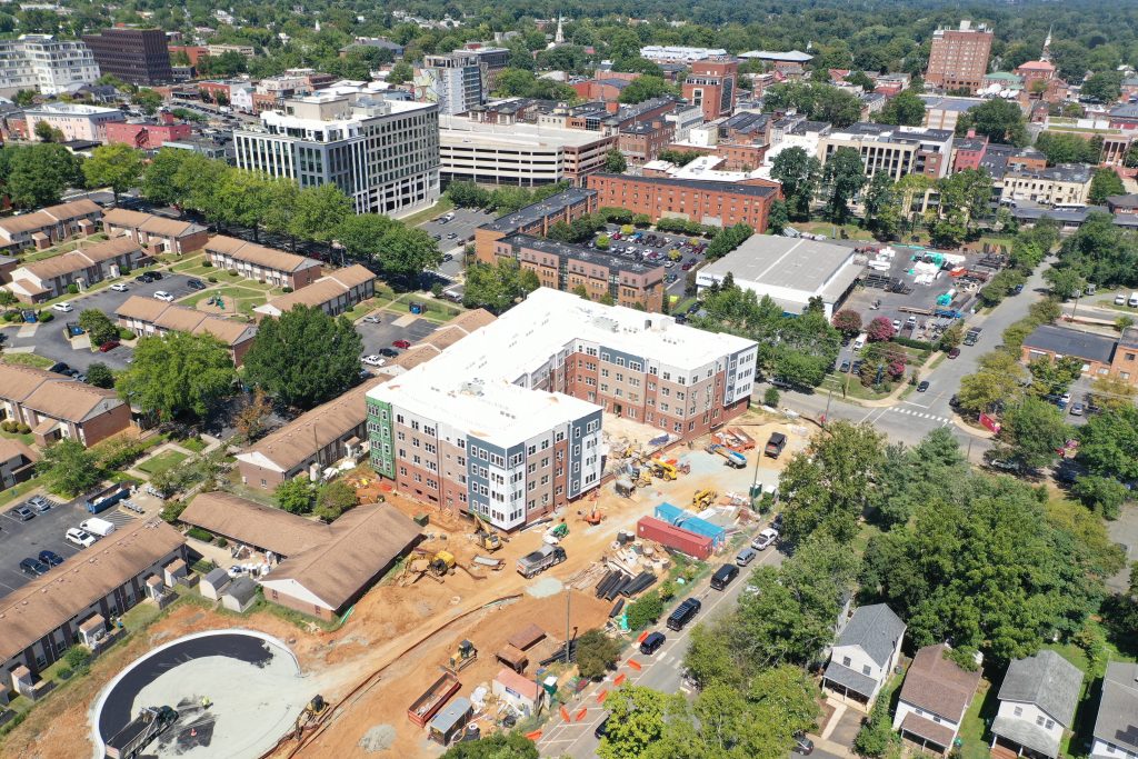 An aerial view of a large, four-story, U-shaped housing development, still being built, and surrounded by settled neighborhoods on the three sides that are visible. The roof is white and the various sections of the exterior walls are blue, tan, brick, or white. The ground around the structure is still raw dirt, with several trucks and machines in view.