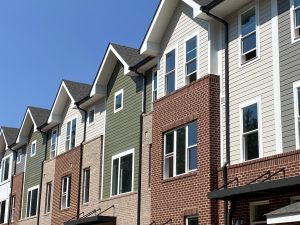 A view from the ground of Kindlewood units. The windows are still blank and devoid of curtains, indicating that the building is still under construction. The narrow, joined apartments have varied fronts and they include tan or red brick, or clapboard, or combinations thereof. The roofs are peaked. The view doesn't include the front doors and there are no people in the photo. The sky is blue.