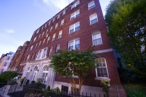 An exterior view of a five-story brick apartment building, with wide steps leading from the sidewalk to the three front doors, over which there are arched windows. There are shrubs and small trees in front of the building and an iron railing. The sky is blue with wispy clouds and there are no people in the photo.