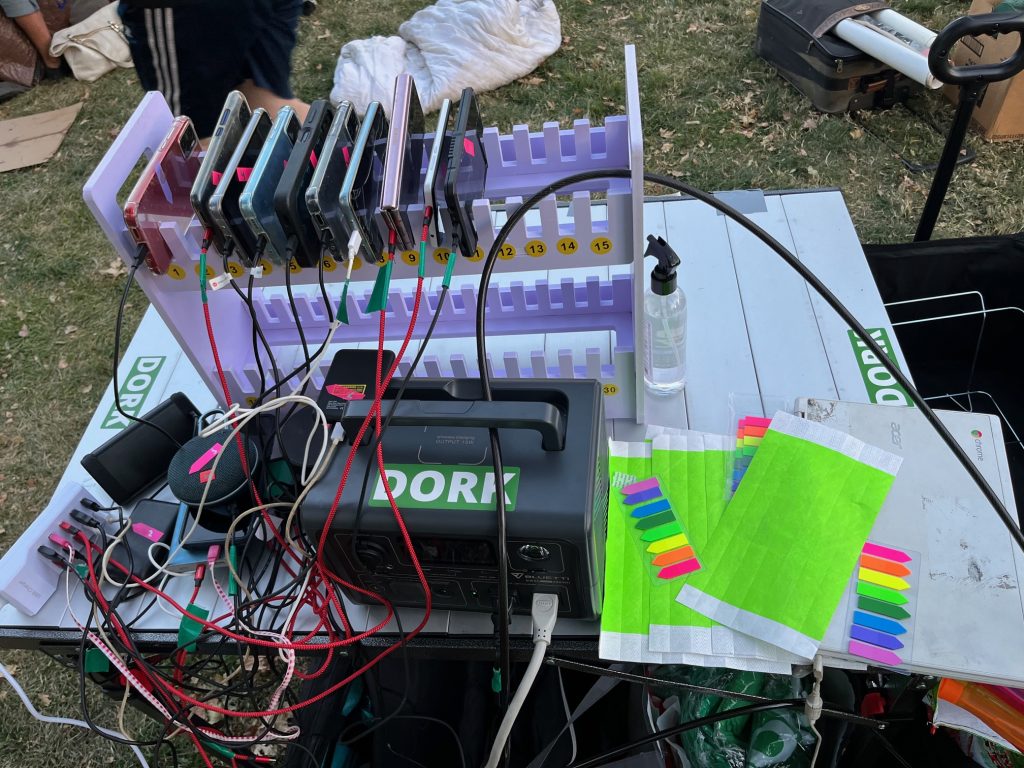 Table or bench with a rack holding 10 cellphones, all plugged into chargers, creating a large tangle of cables A large battery or charger has a sticker that says, in white capitals on green, DORK. Also on the table are multicolored sticky notes.