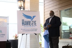 An easel holds a square poster that reads "Hawk's Eye Apartments/Brought to you by CareOregon and Columbia Pacific CCO. Next to the easel stands a tall Black man with a salt-and-pepper beard, wearing a black suit over a plaid shirt. He's holding a blue banner with lettering but the fabric is bunched up and the lettering can't be read.