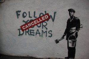 A mural on a rough whitewashed wall. Painted in dripping capitals is "Follow your dreams," and over it is a red stenciled "Cancelled" banner. To the right is an image of a man holding a pail and paintbrush. He's wearing a cloth cap and has two rolls of paper under his arm.