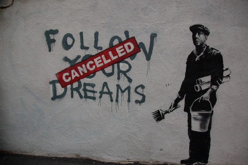 A mural on a rough whitewashed wall. Painted in dripping capitals is "Follow your dreams," and over it is a red stenciled "Cancelled" banner. To the right is an image of a man holding a pail and paintbrush. He's wearing a cloth cap and has two rolls of paper under his arm.