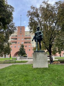 A statue of a man in classical garb stands on a lawn in front of a plaza leading to a very large apartment building in pink and beige tones. Behind the statue on the lawn are two leafy trees. High on the building, barely visible behind the foliage, are the letters "Karl Ma..." (for Karl Marx Hof).