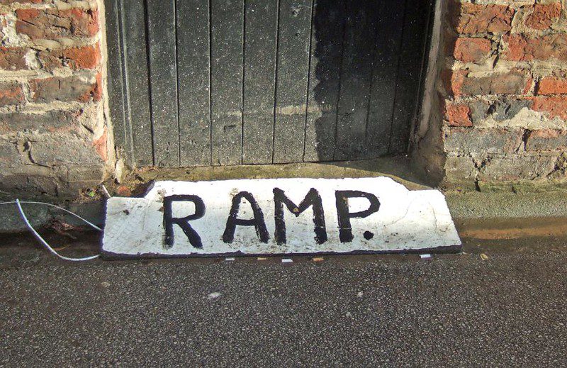A small homemade ramp made of a white board with "RAMP" painted on it in black covers the gap in a street-level doorway of a brick building. The door is of vertical black boards and is dirty with dust and splashed mud.