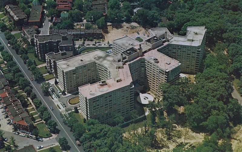 Aerial shot of a huge hotel, 12 or 13 stories high, surrounded by mature trees, other apartment buildings or hotels, with a roadway in front of it. The building is shaped vaguely like a stick figure of a person, but with a C-shaped head.