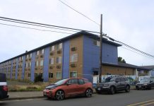 A long three-story building, a former motel, painted in medium brown and blue wide verticals, seen from one end. A chain-link construction fence blocks access to the building from the sidewalk. Three cars are parked on the curb by it. There are no people in the photo.