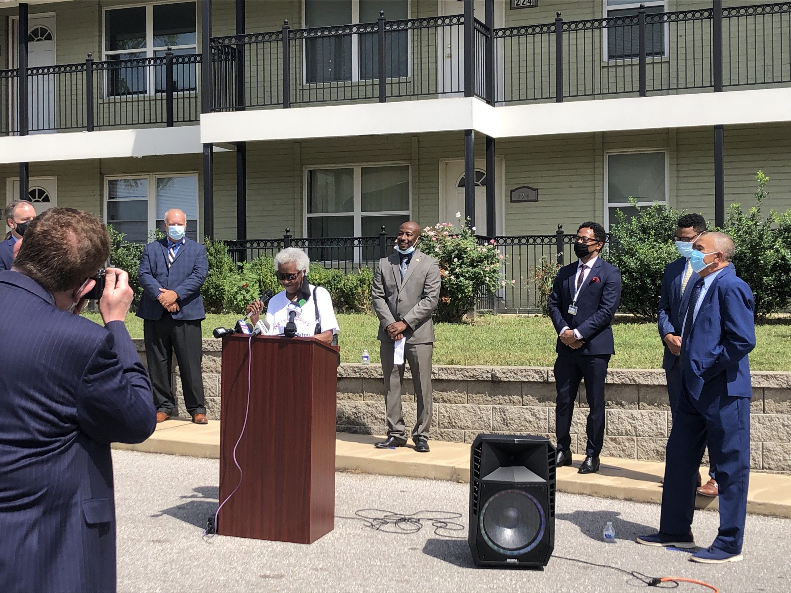 A Black woman with white hair stands at a podium in the parking lot of a residential building (in Wellston, Missouri). She is wearing sunglasses and smiling. Behind here are six men of varying ages, all in suits, most wearing masks. In the foreground is a back view of a photographer.