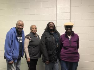 Four smiling middle-aged African-Americans standing in a row in a hallway, with a cream-painted cinderblock wall behind them. From left, a white-haired man in a blue zip-up hoodie, a woman with short white hair and wearing a black fleece vest, a long-haired woman in a black parka, and a woman with white hair in a purple fleece top and a cloth cap. Her face is in shadow but you can see her smiling.