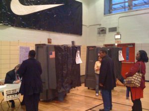 A polling place in a space that appears to be a gymnasium. (A large Nike "whoosh" banner hangs high on the wall.) There are two curtained voting booths on wheels, and the lower legs and feet of a voter can be seen behind one of the curtains. The second booth is partly hidden, but a woman in jeans and a blouse is standing in front of it. She may be a voter or a poll worker. A volunteer poll worker seated at a folding table is signing in a voter, and two other people wait in line to sign in.