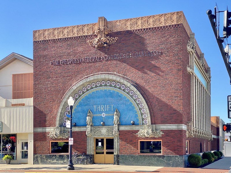 View from the street of a bank built in 1917. From the photographer: The building features a red brick exterior with terra cotta trim, decorative panels with Sullivanesque detailing, Sullivanesque trim, a decorative mosaic in the tympanum below the arch above the front entrance with the word “Thrift” in gold lettering in the middle of an expanse of blue tile and decorative white, cream, green, purple, red, and orange tile accents, decorative metal lettering on the facade above the arch displaying the words “The People’s Federal Savings & Loan Assn." ... Gargoyles above the pilasters framing the front entrance, fixed glass windows at the corners, brass double doors.