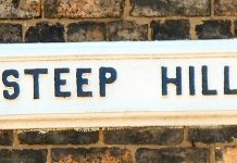 A sign on a brick wall advising drivers of a steep hill. The sign is all-caps black lettering on a white background.