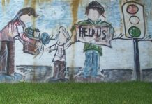 A large graffiti'd mural, painted in a cartoonish style, showing an apparently homeless family of three standing near a traffic light. The mother is handing a box or carton of stuff to the child, whose other hand is reaching to the father figure. He is holding up a sign that says "Help us." Facial features were not drawn on these figures, giving them a universal quality.