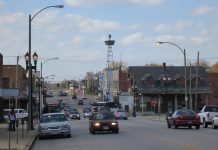 A streetscape of a town on a partly cloudy day. Cars travel the main road toward and away from the camera. In the middle distance is a tall radio tower. Identifiable businesses include a laundromat and beauty supply store.