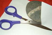 Close up view of rock, scissors, and a sheet of white paper on a red table or desk. The rock is oval gray rock with a band of white quartz through it, the scissors have purple plastic grips and metal blades. The scissors are positioned such that they appear to have just cut the paper, and the rock is resting on the paper, half concealed by a portion of it.