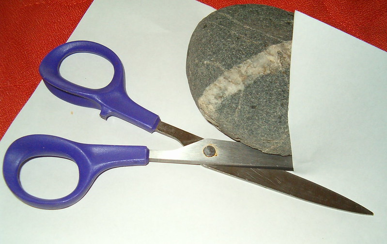 Close up view of rock, scissors, and a sheet of white paper on a red table or desk. The rock is oval gray rock with a band of white quartz through it, the scissors have purple plastic grips and metal blades. The scissors are positioned such that they appear to have just cut the paper, and the rock is resting on the paper, half concealed by a portion of it.