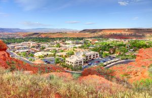 St. George, Utah, seen from a hillside. In the foreground are deep brick-red and orange rocks, with light green/yellow grasses. The town lies in a valley, and trees lining the streets are a darker green than the grasses. Far in the distance are mountains, under a blue sky with sparse clouds.