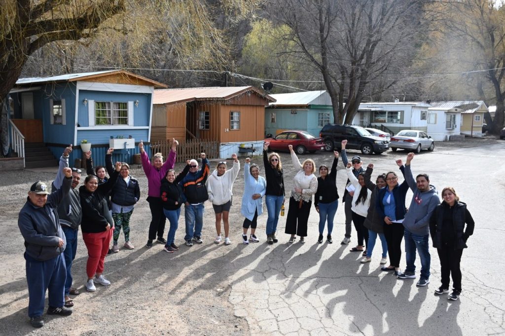 Nineteen people stand in a semi-circle facing the camera, on a partly paved road near some mobile homes and parked cars. They are mixed in age, gender, and skin color, and most have one arm raised high, making a fist; one person is raising both arms with thumbs up, and one person has not raised her arms.