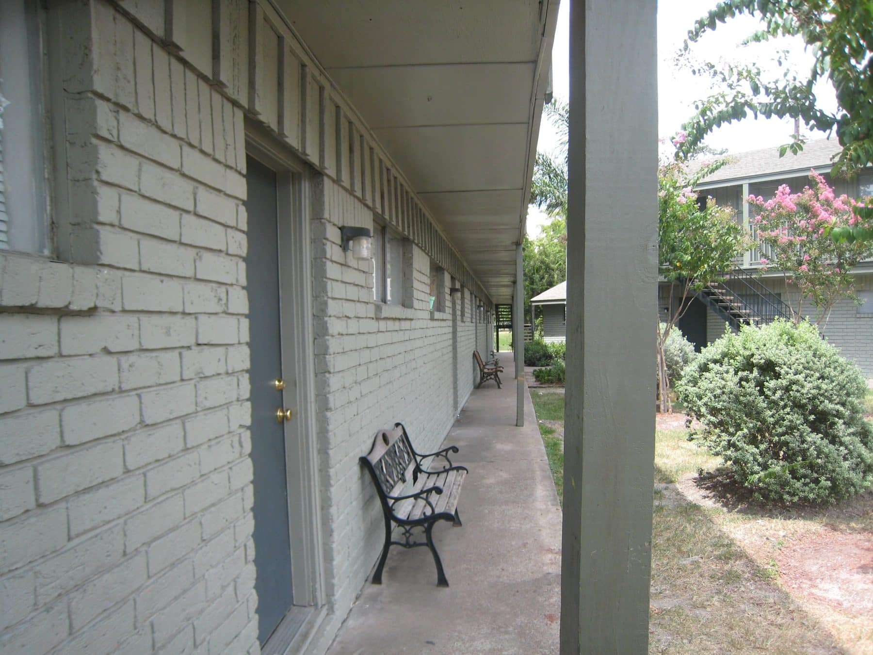 View from one end of the front of a long motel-like apartment building. The brick front is painted a sage green, and a roof overhangs the concrete walkway. Slatted wooden benches are placed between the front doors, which are a darker green than the building. In front of the building are shrubs including crape myrtle, and on the other side of the small yard can be seen part of a similar-looking building.