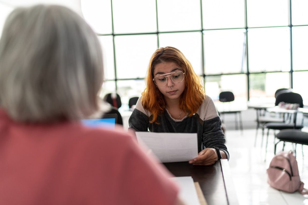 A young woman in gold-rimmed glasses looks skeptically at a sheet of paper. She is across a table from an older woman in a peach top whose back is to the camera. The young woman has wavy brown hair with red overtones and wears a top that's light on top and dark below the shoulders. They're in a brightly lit meeting room.