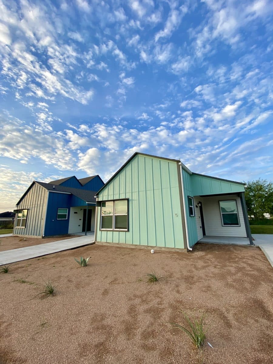 A small green house on a lot not yet planted with grass or shrubs. Vertical siding is light green, and the entryway at the right is overhung by a roof. At left is a similarly designed house in dark blue. Behind the houses the bright blue sky is dotted with cirrus clouds.