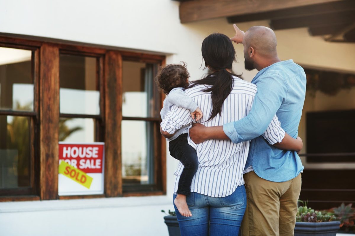 A young family of three seen from the back as they look at a house. From right: A light brown-skinned man with shaved head and chin whiskers in a blue chambray shirt and khakis points to the house, at something out of frame. His other arm is around a black-haired woman in a narrow-striped button-up white shirt over blue jeans. One of her arms is around the man's waist; with the other she holds a small dark-haired child in a pale blue top and black leggings and no shoes. The house is white with brown window trim, and a sold sign in one window.