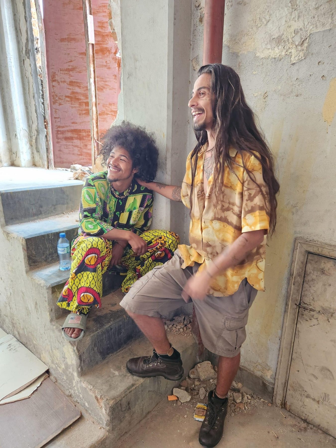 Two men, one with long hair and his hand on the shoulder of the other man, who is sitting. Both are smiling. The man on the right is wearing a yellow shirt and shorts. The man on the left is wearing a patterned green shirt and pants.