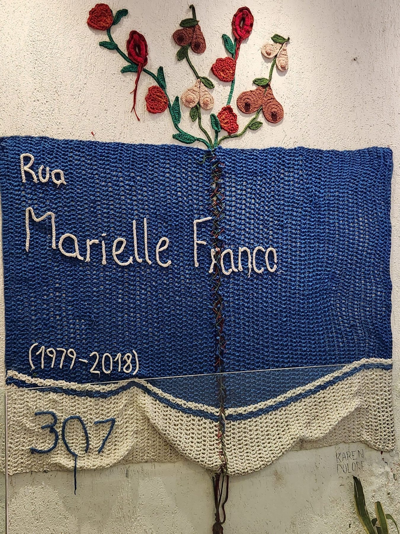 A blue and white crocheted square reads "Rua Marielle Franco (1979-2018), 307." Crochet flowers extend from the top.