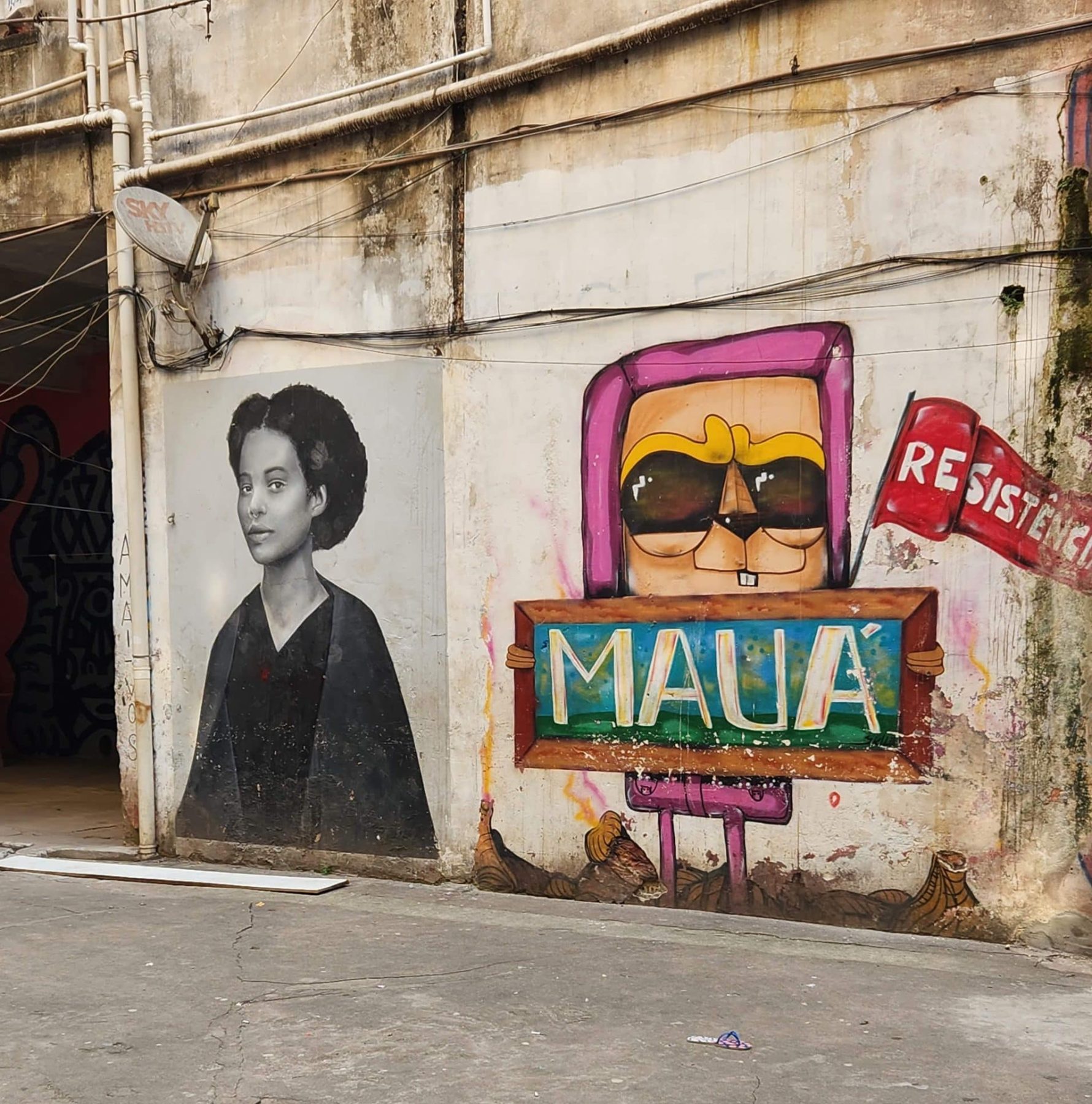 A small black and white mural of a woman in robes, next to a small mural of a cartoon animal holding a sign that says "Mauá" and a flag that says "resistência"