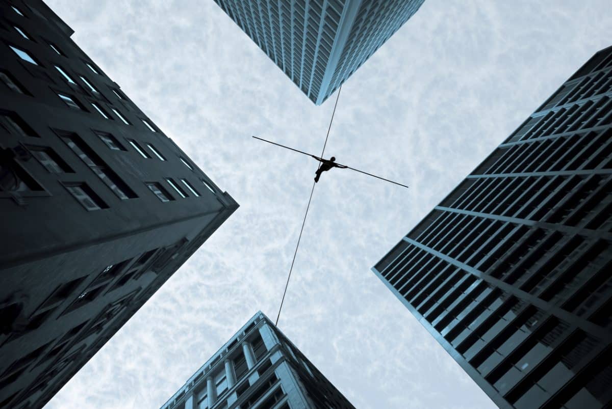 A tightrope walker pictured from below walking across a line between buildings.