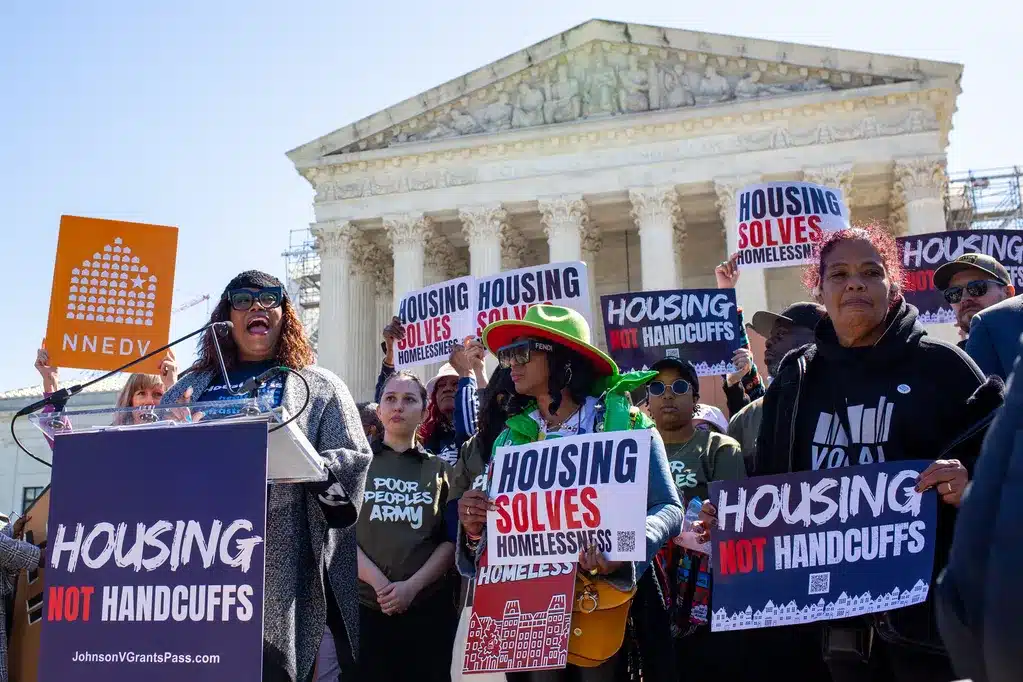 A crowd of protesters gather in front of the U.S. supreme court. One woman is speaking. They are holding signs that say "housing solves homelessness" and "housing not handcuffs."