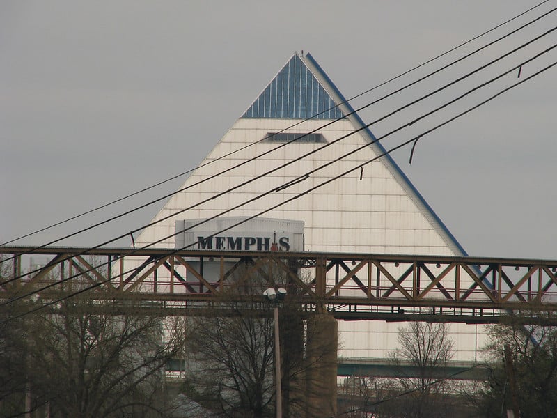 A pyramid-shaped building, mostly white but blue at the top. In front of it is a tall sign that reads "Memphis" in capital letters, with a guitar standing in for the letter I. In front of that is a trestle of brownish metal, and crossing the view diagonally are five parallel power lines.