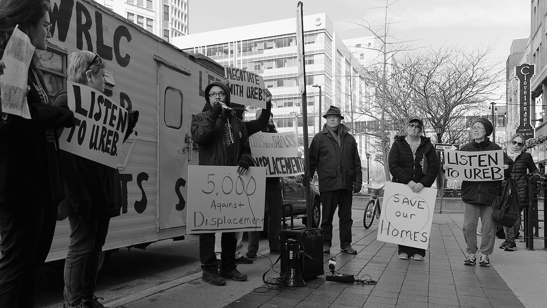 Seven people wearing jackets and caps on a city sidewalk holding signs that say "Listen to UREB," "Save Our Homes," "Negotiate with UREB," or "5,000 Against Displacement." One person is speaking into a microphone. At the curb by the speaker is a van with WRLC painted on the side, for Western Reserve Land Conservancy.