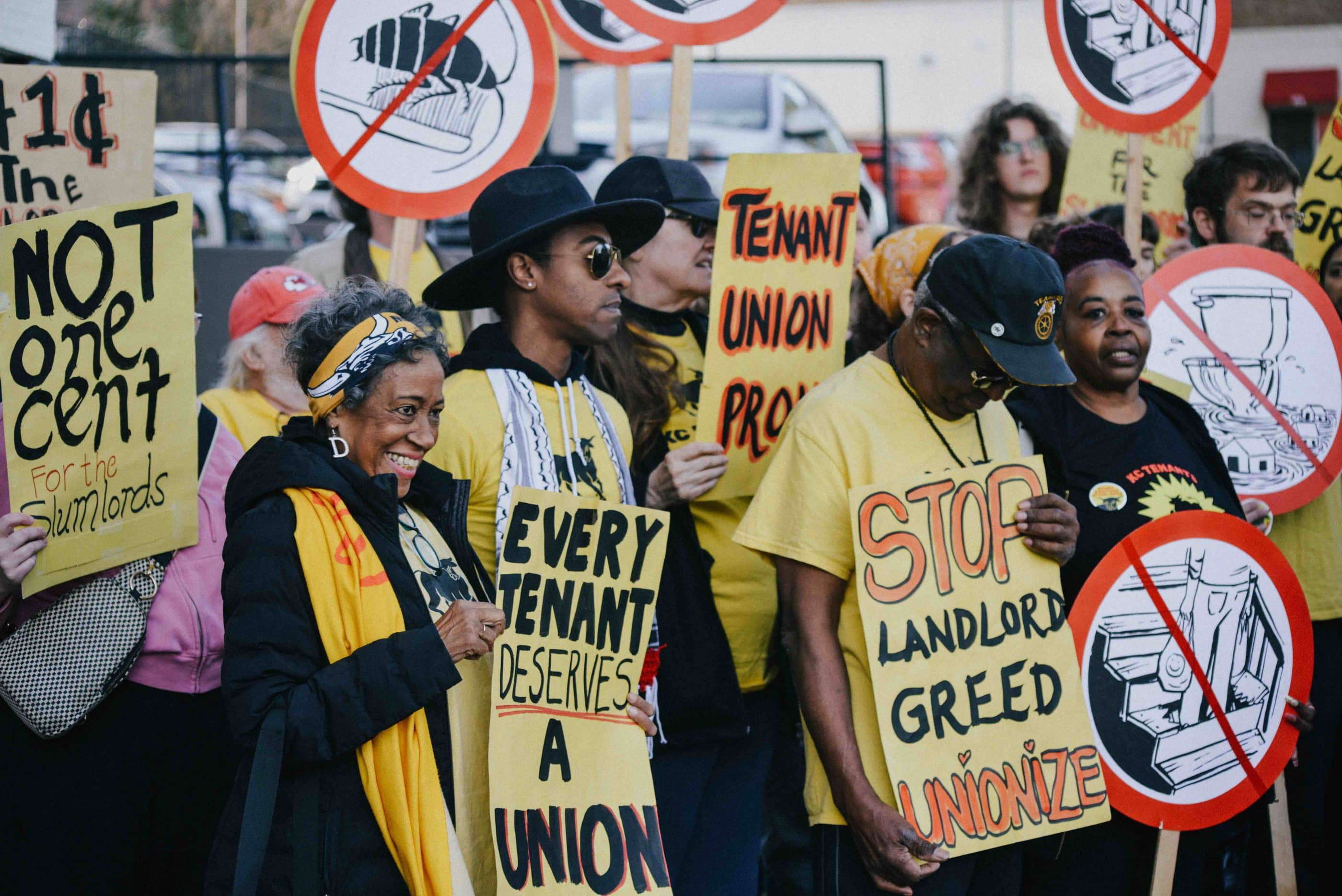 Busy scene of striking tenants, of mixed ages and skin tones, most holding signs. Signs say "Stop landlord greed/Unionize" and "Every tenant deserves a union" and "Not one cent for the slumlords." Others are round "universal no" signs showing rodents, broken staircases, flooded bathrooms.