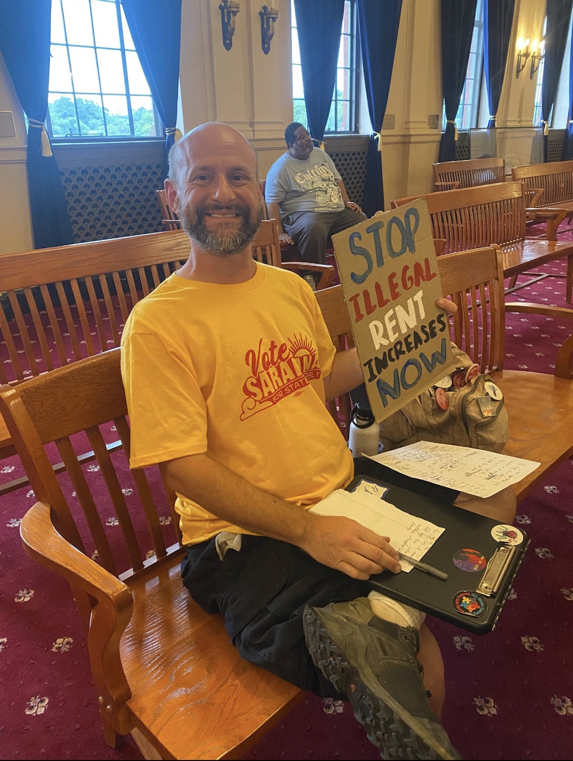 A balding, bearded man in a bright yellow T-shirt is seated on a wooden chair in a public meeting room. His shirt reads "Vote Saranna" and he holds up a hand-done poster that reads "Stop Illegal Rent Increases Now."