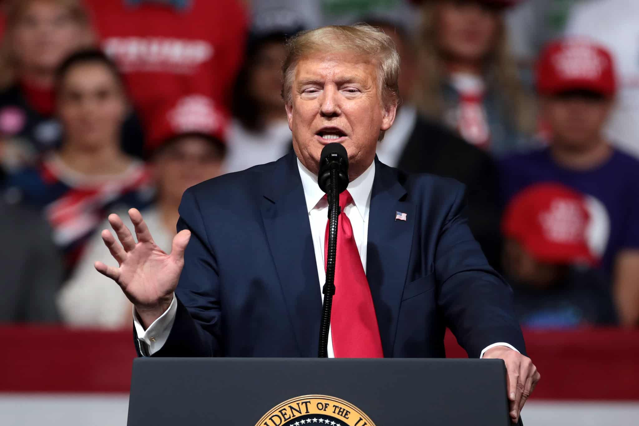 A white man with blond hair, a blue jacket, white shirt and red tie stands in front of a blue lectern that has the presidential seal on it. The man is gesturing with his right hand as he speaks into a microphone.