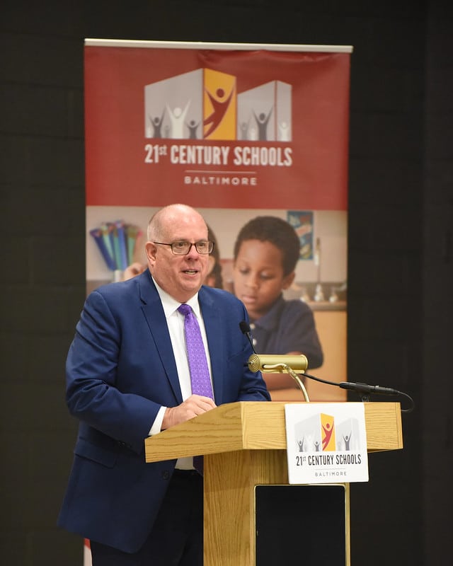 A middle-aged white man in a blue suit and purple tie speaks at a lectern. Behind him is a large banner with the heading "21st Century Schools/Baltimore." The man speaking is covering one of the faces in the banner, but also in the image is a small Black boy, giving the momentary impression that he is standing next to the man at the microphone.