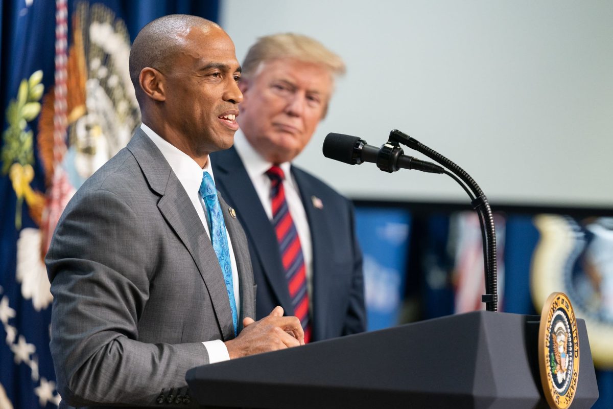 Scott Turner, a shaven-headed Black man, seen in partial profile at a lectern. Wearing a gray suit, white shirt, blue tie. Beyond him and out of focus is Donald Trump.