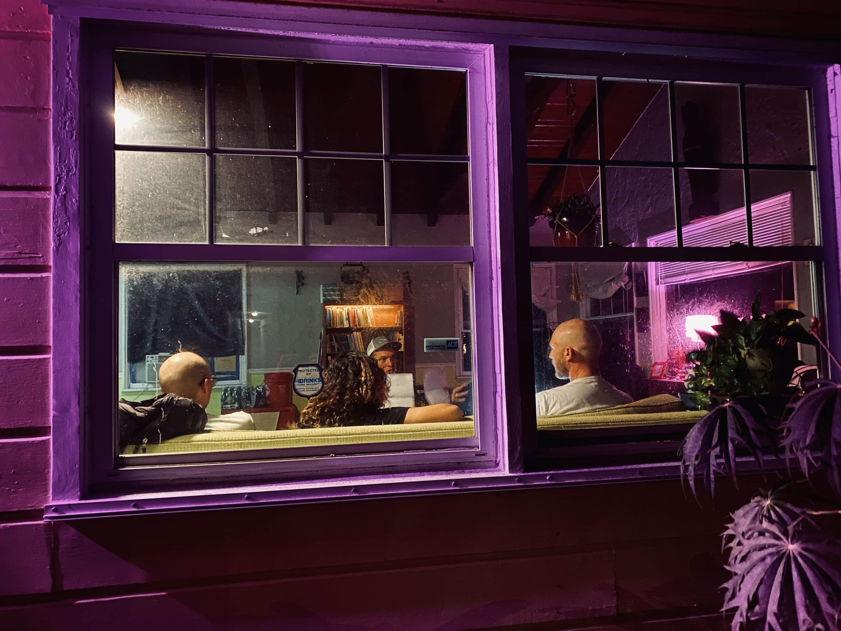 Looking through a purple-framed window to a nighttime meeting of four people. Three with back to camera, two bald white men and a woman with curly dark hair. Facing the window, a white man in a ball cap is speaking.