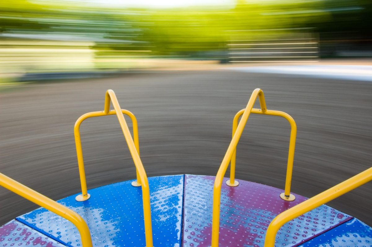 View from the center of a spinning playground merry-go-round. It's in focus but the background is quite blurry, in shades of green with a gray ground . The metal platform was painted blue and red but is somewhat worn, showing bare metal, and the handholds are painted bright yellow. No children are in the photo.