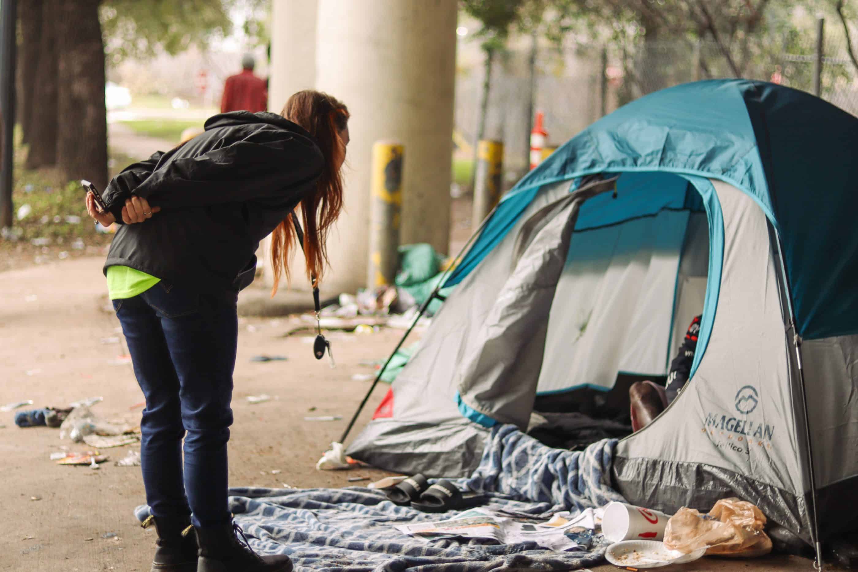 A young woman leans forward to address the occupant of a dome tent. The person in the tent is mostly hidden except for a knee. On the ground around the tent are food wrappers, slippers, and a newspaper.