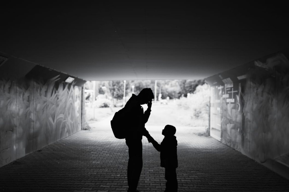 Black and white image showing parent and child silhouetted in a tunnel. Adult has a backpack and is holding one hand to their forehead, conveying worry or anxiety. Child is holding the adult's other hand and looking up. Tunnel appears to be an underpass, far end is blurry but looks like grass and greenery.