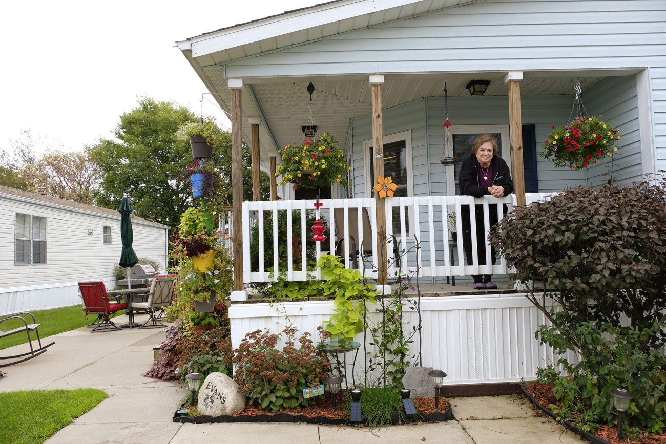 A smiling middle-aged white woman in a black jacket leans over the white porch railing of a blue house surrounded by shrubs and plants. On either side of her are hanging pots of colorful flowers. To the left of the house is a round patio table with furled umbrella and four chairs.