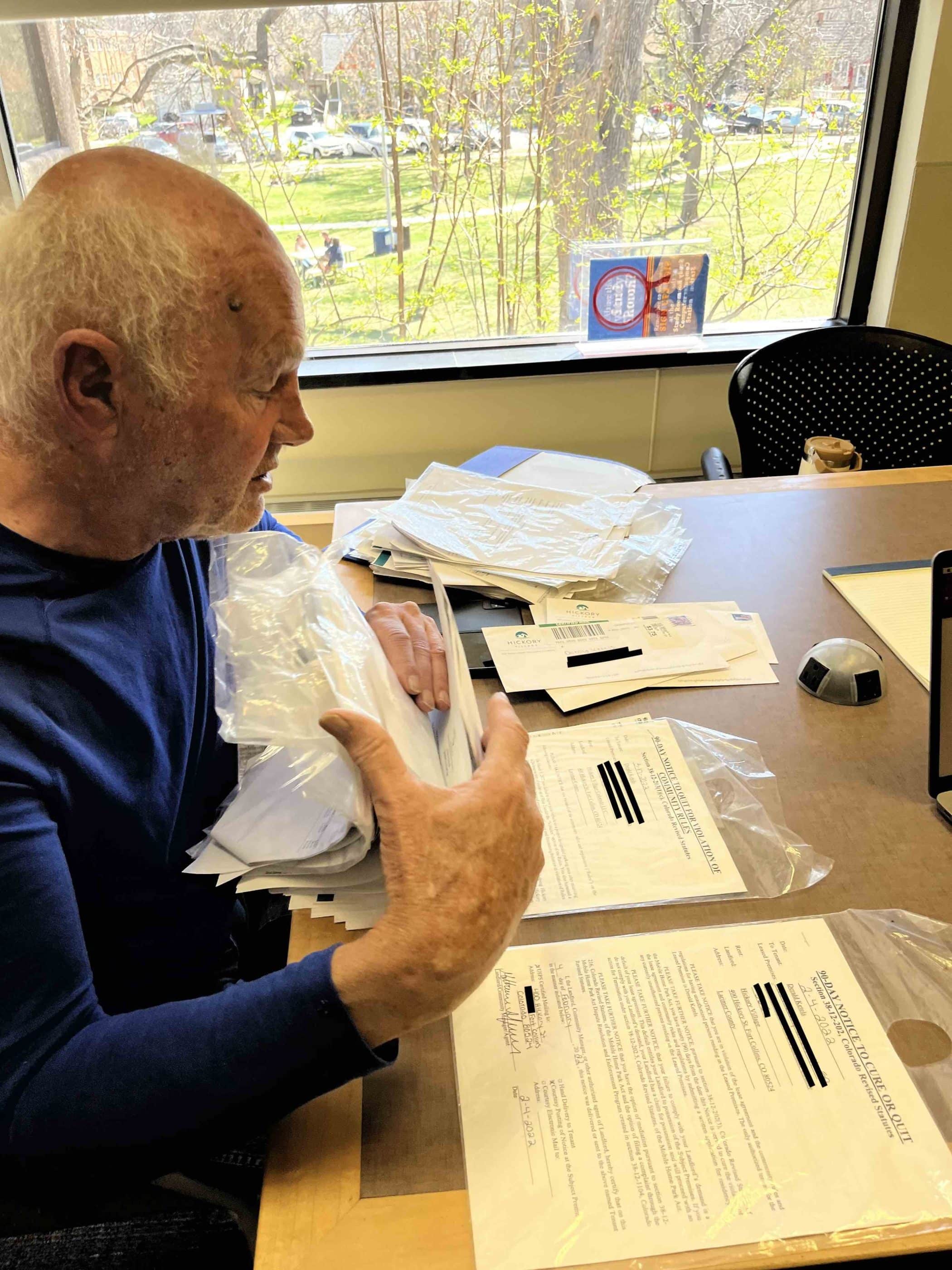 An elderly white man in a blue knit shirt seen from the side sits at a table spread with documents.