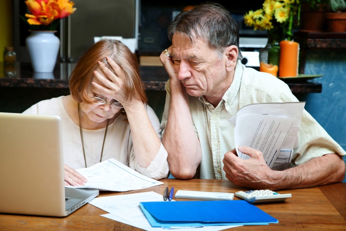 An older white couple sits side by side at a table covered with paper, folders, calculator, pens, and laptop. They're looking at a document with apparent worry or anxiety.