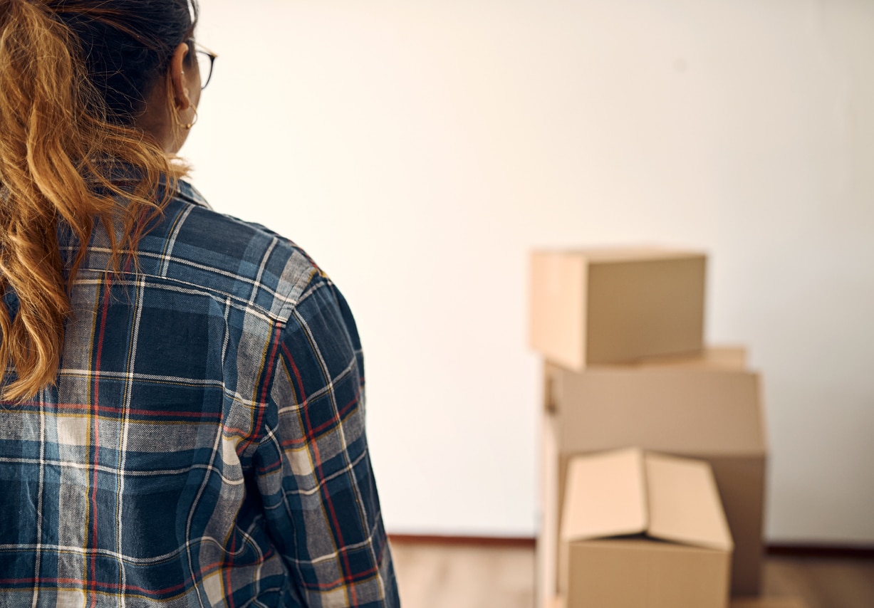 Rear view of a pony-tailed youngish woman in a plaid shirt and wearing hoop earrings facing a blank wall in front of which is a pile of moving boxes, not quite in focus.