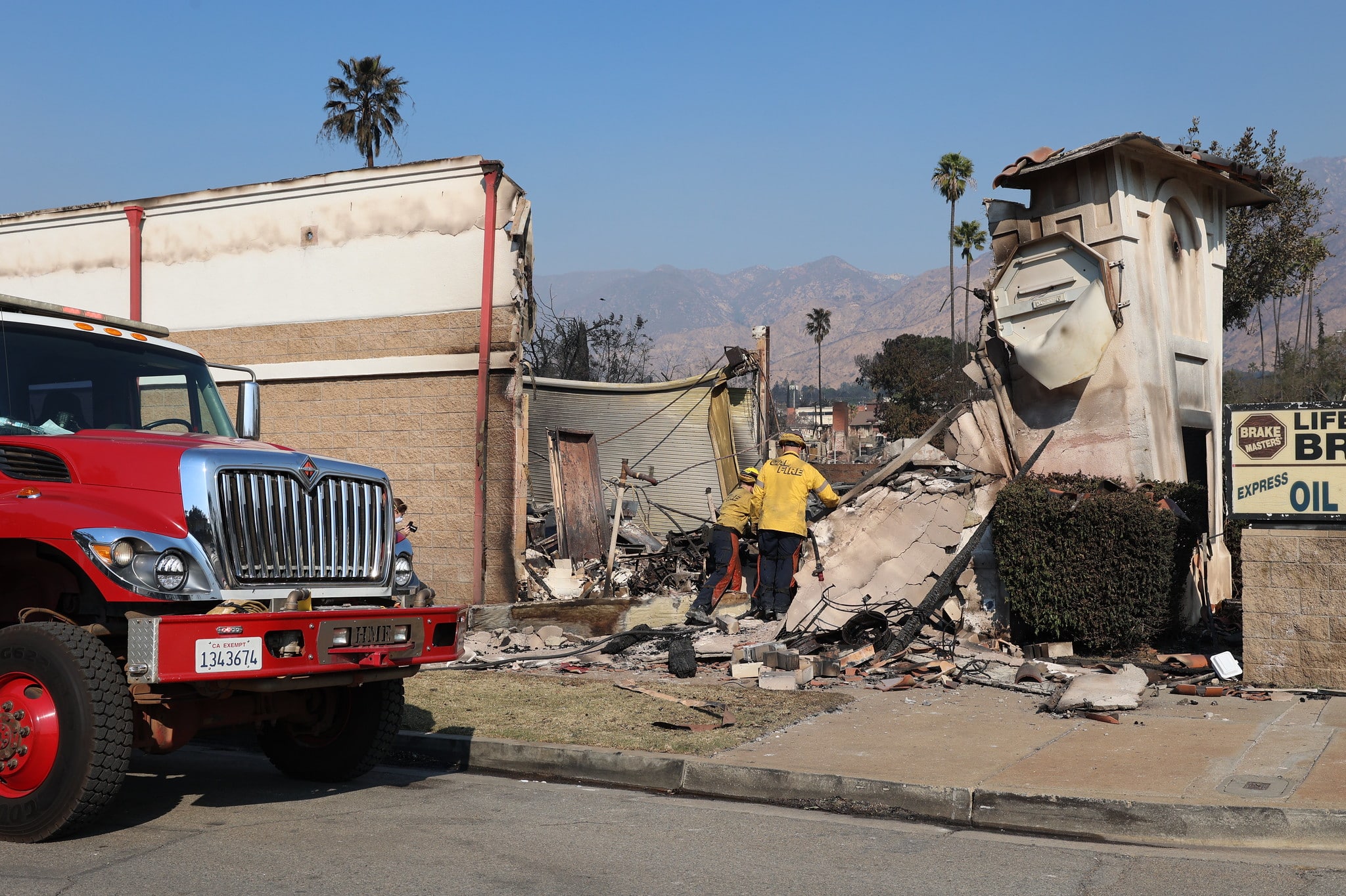 There is a firetruck on the left and two firefighters wearing yellow entering a badly burnt property. In the background are hills and palm trees.