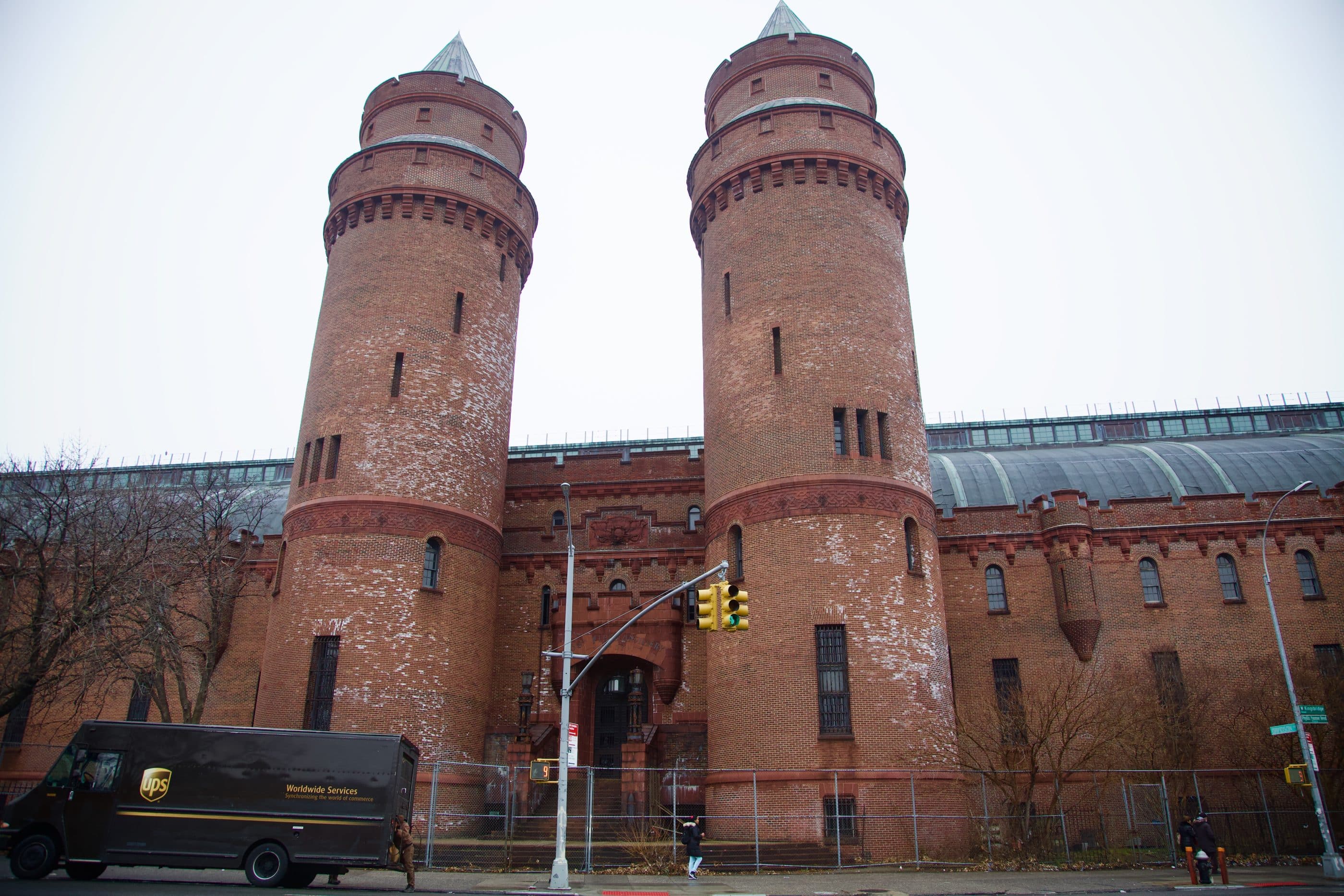 A massive 9-story red brick armory with a curved metal roof fills the frame from left to right. Reminiscent of medieval architecture, the edifice has two tall crenelated towers with conical roofs flanking the main entrance. A chain-link fence borders the property.