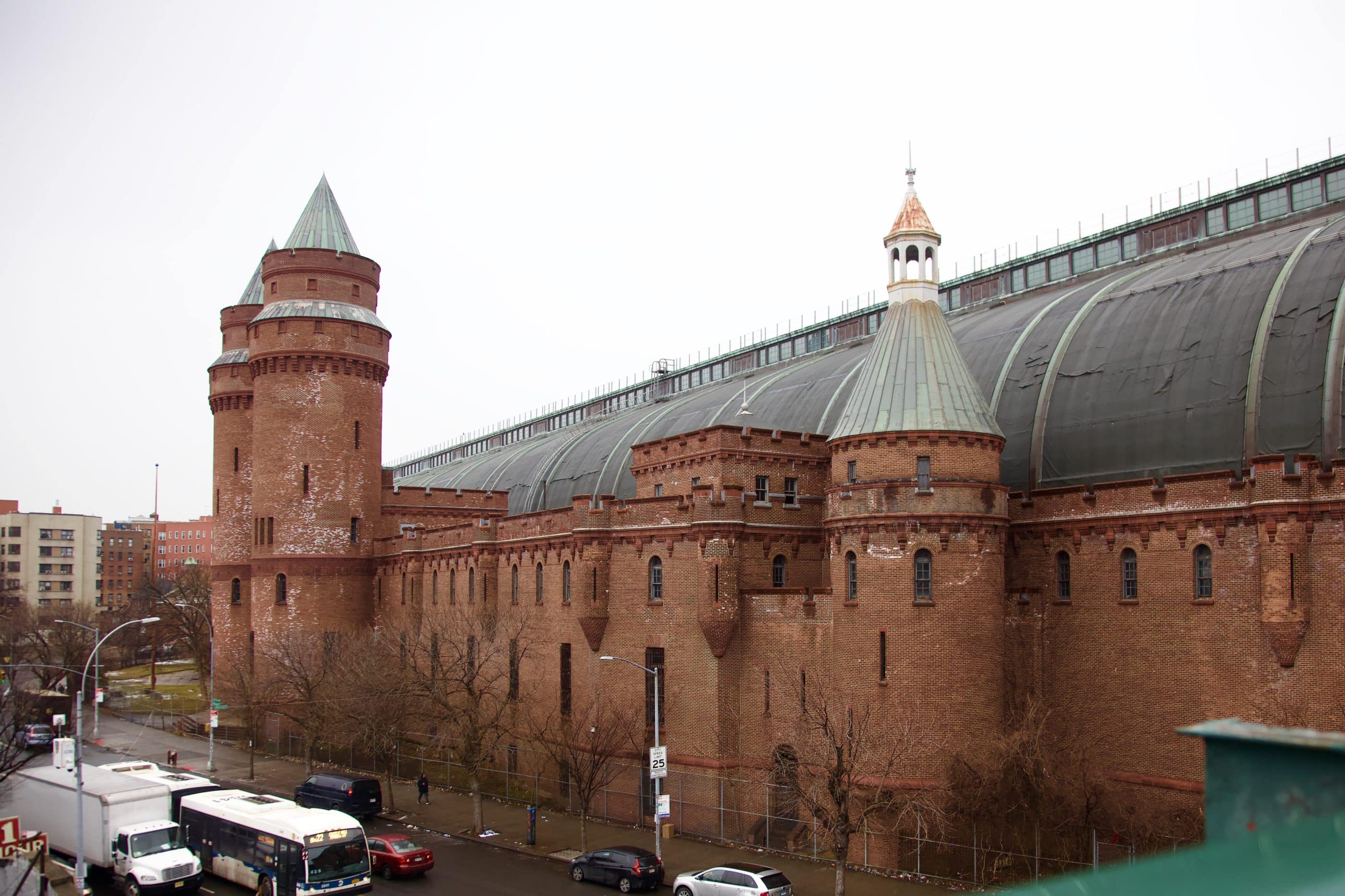 A massive 9-story red brick armory with a curved metal roof, seen from one end. Reminiscent of medieval architecture, the edifice has two tall crenelated towers with conical roofs flanking the main entrance, and another, shorter tower topped by a gazebo. A chain-link fence borders the property, and buses, trucks, and cars can be seen in the street, and pedestrians on the sidewalk.