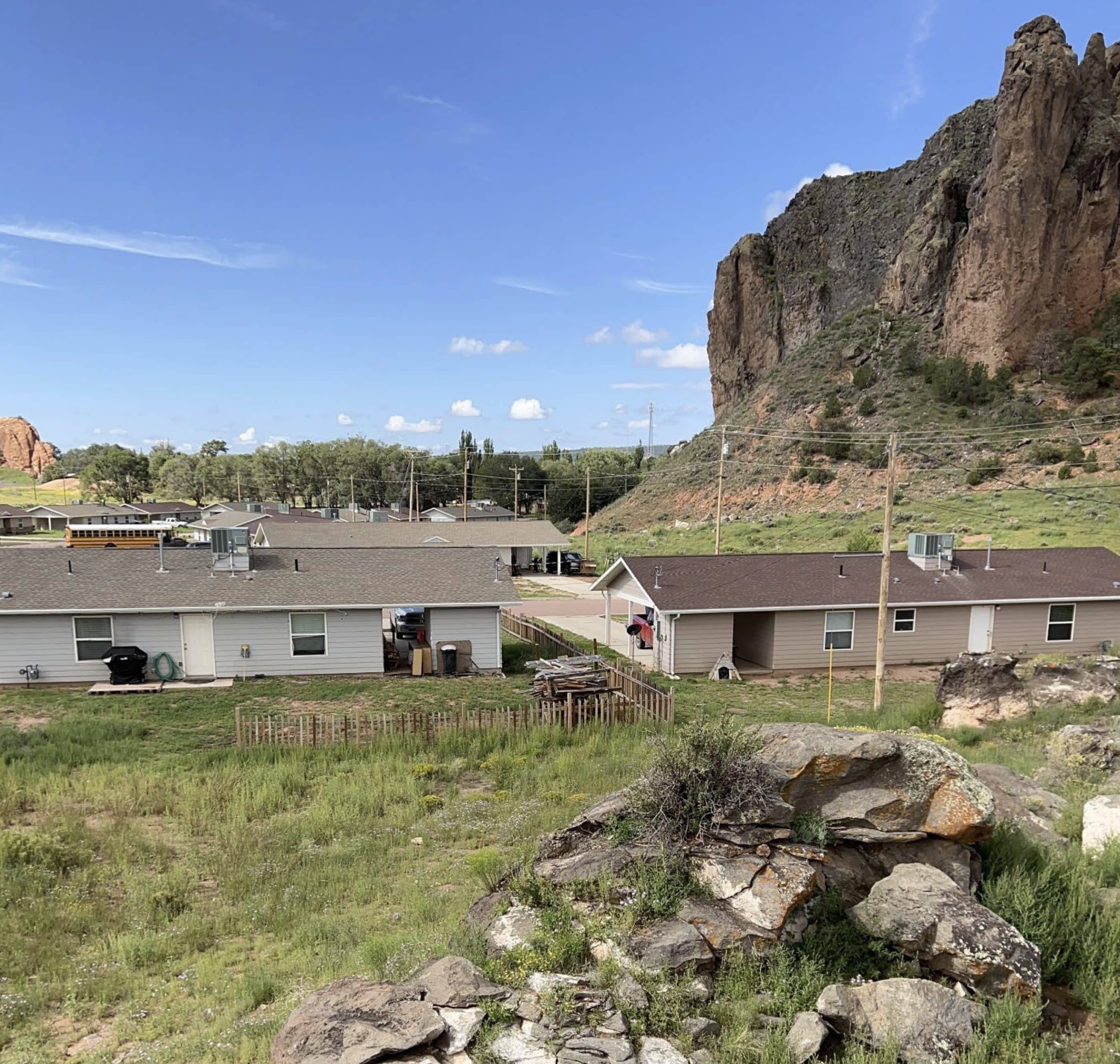 Landscape under a blue sky, with large looming rock cliff at right, single-story housing in the middle distance, and grass and rocks in the foreground. In the far distance is another rock formation.